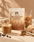 A bag of Mataye Mushroom Coffee on a tan surface next to a mug of latte and a glass of iced mushroom coffee, styled with whole lions mane and reishi mushrooms, coffee beans, and a gold measuring scoop of cocoa powder.