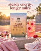 A bag of Mataye Super Yerba Mate is displayed on a concrete track surface at sunset, styled with a pink drink of raspberry lemonade, pink running shoes, sunglasses, and a pink towel. The background shows a blurred running track and field.